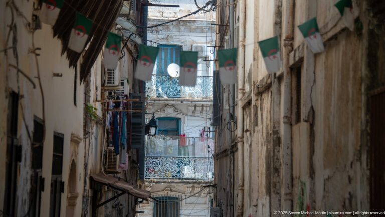 Narrow Casbah alley with Algerian flags, wrought-iron balconies, shutters, laundry, satellite dishes, and overhead wires.