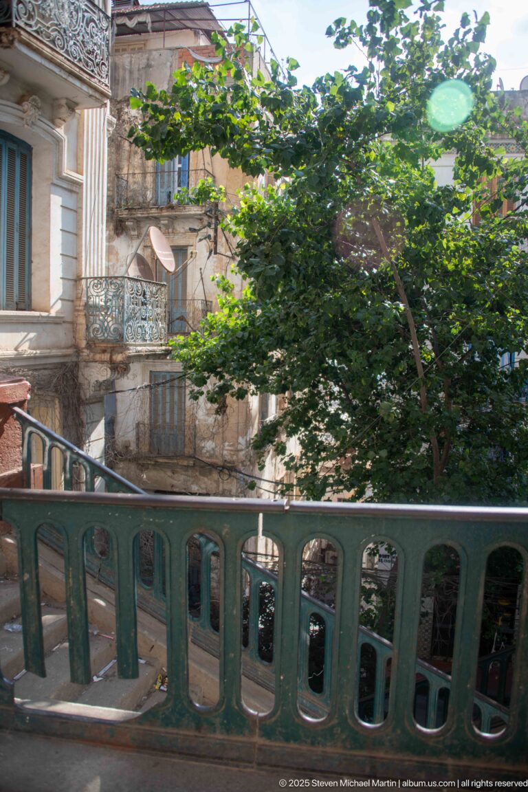 Green balustrade in front of dense trees and a weathered residential building with balconies and satellite dishes.