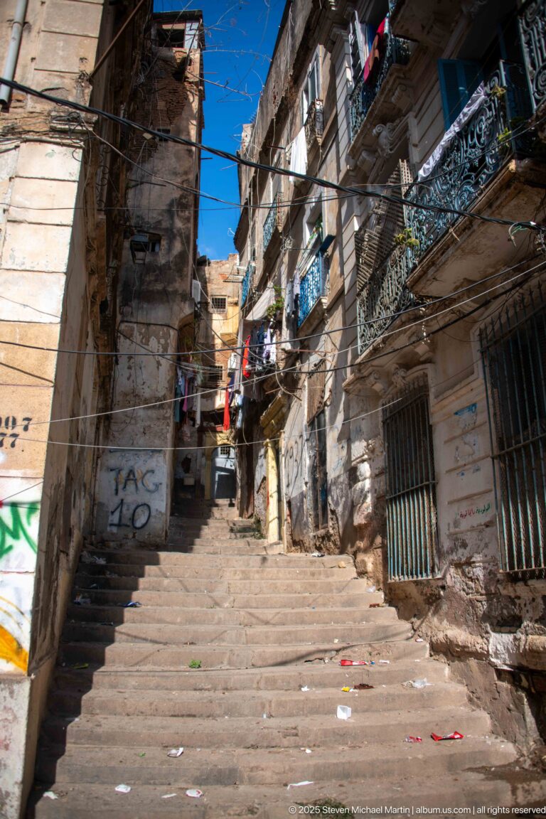 Long stone stairway climbing between dense Casbah residences with balconies, laundry, barred windows, and overhead wires.
