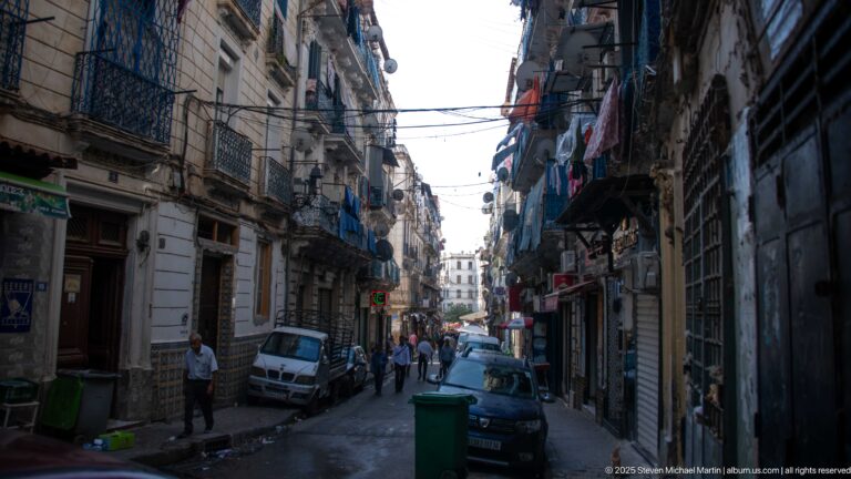 Narrow busy street with pedestrians, parked cars, shops, balconies, hanging laundry, and dense overhead wires.