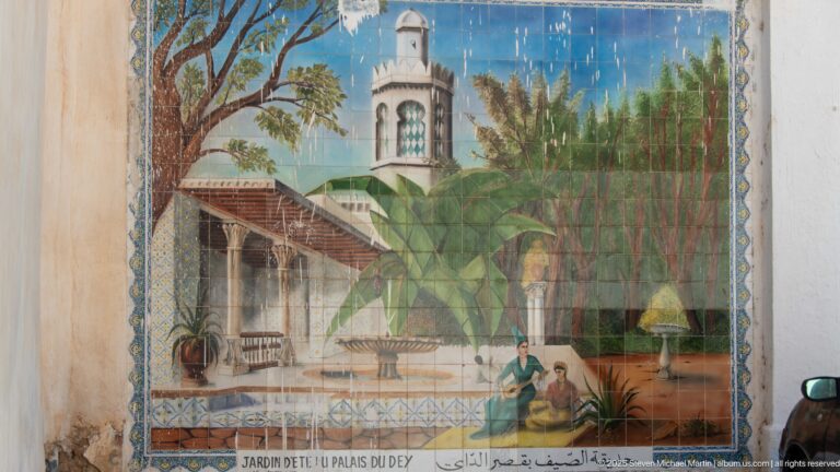 Weathered decorative panel showing a palace garden with palms, fountain, figures, architecture, and French and Arabic text.
