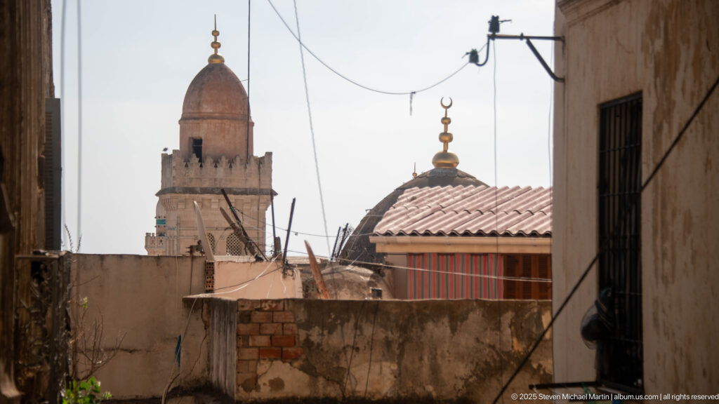 Mosque dome with crescent finial and tall masonry tower rising beyond weathered Casbah rooftops and overhead wires.