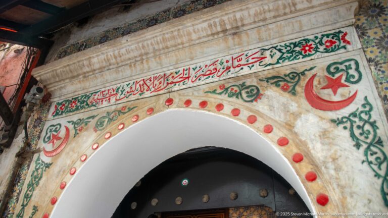 Arched entrance decorated with Arabic writing, floral designs, red dots, crescent-and-star motifs, and surrounding tilework.