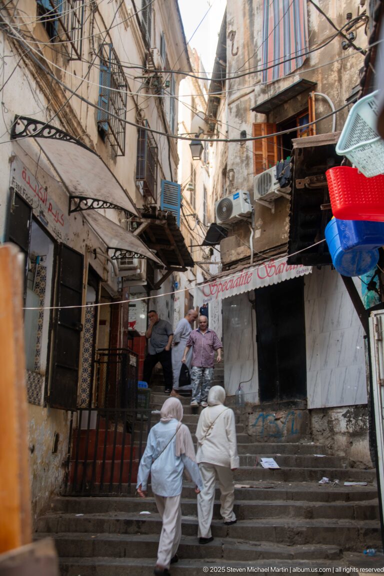 People gathered on a stepped market lane lined with shops, awnings, baskets, and overhead cables.