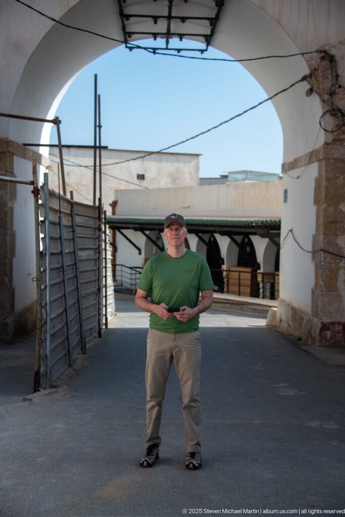 Visitor in green shirt standing beneath a broad white arch amid weathered walls and shaded openings in the Casbah.