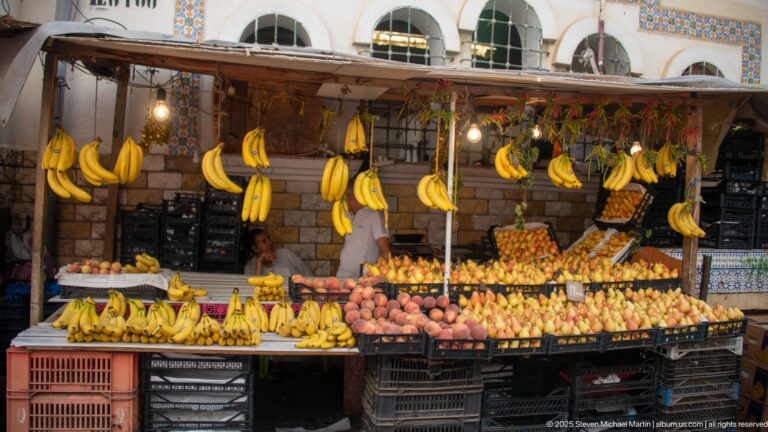 Fruit stall with many hanging banana bunches and crates of mixed produce beneath a tiled building façade.