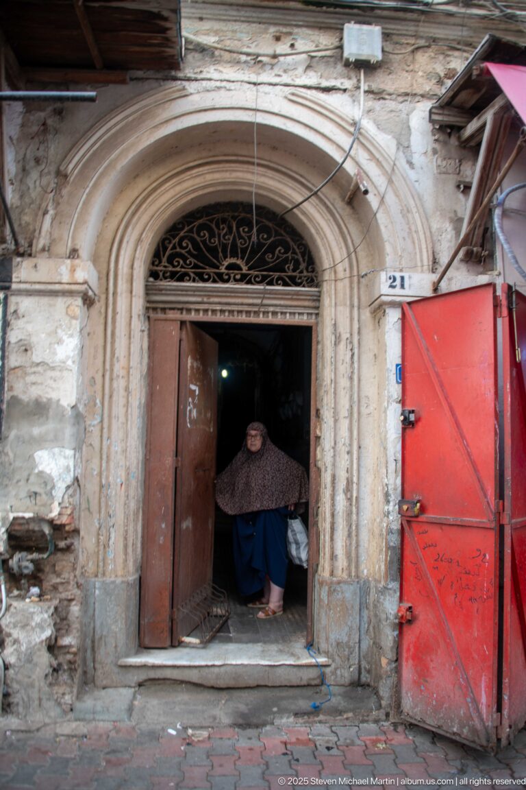 Woman seated inside a tall arched doorway with carved stone surround, wrought-iron transom, and bright red shutters beside it.