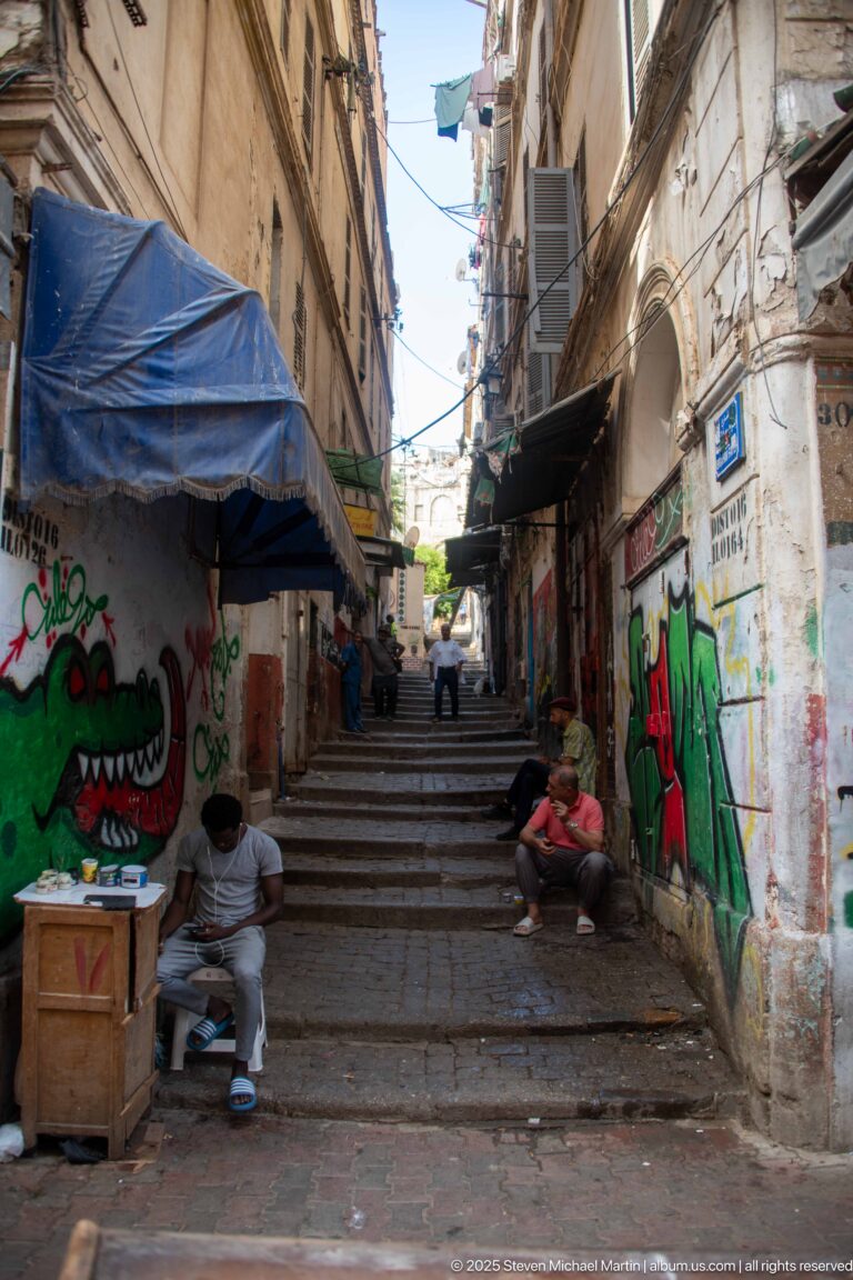 Stepped Casbah lane with colorful graffiti, shop awnings, seated men, and narrow buildings.