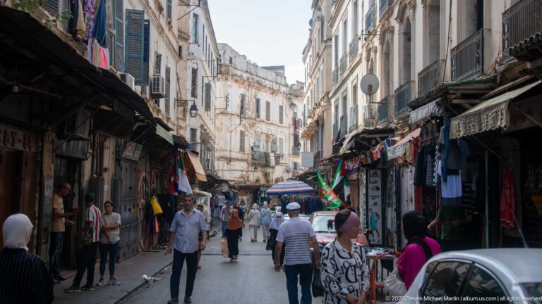 Busy pedestrian shopping street in the Casbah with shops, hanging clothes, awnings, vehicles, and white multi-story buildings.