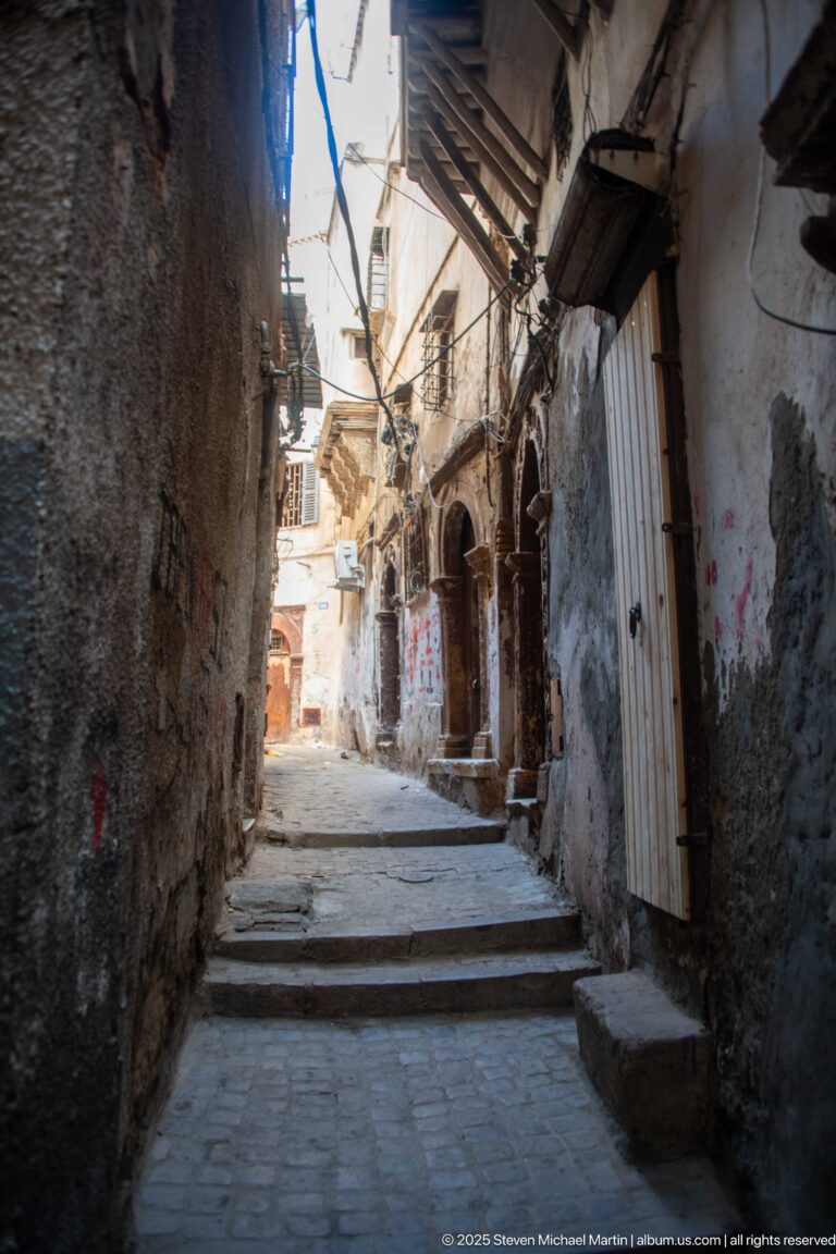 Narrow Casbah alley with shallow steps, rough walls, arched doorways, cables, and bright light at the far end.