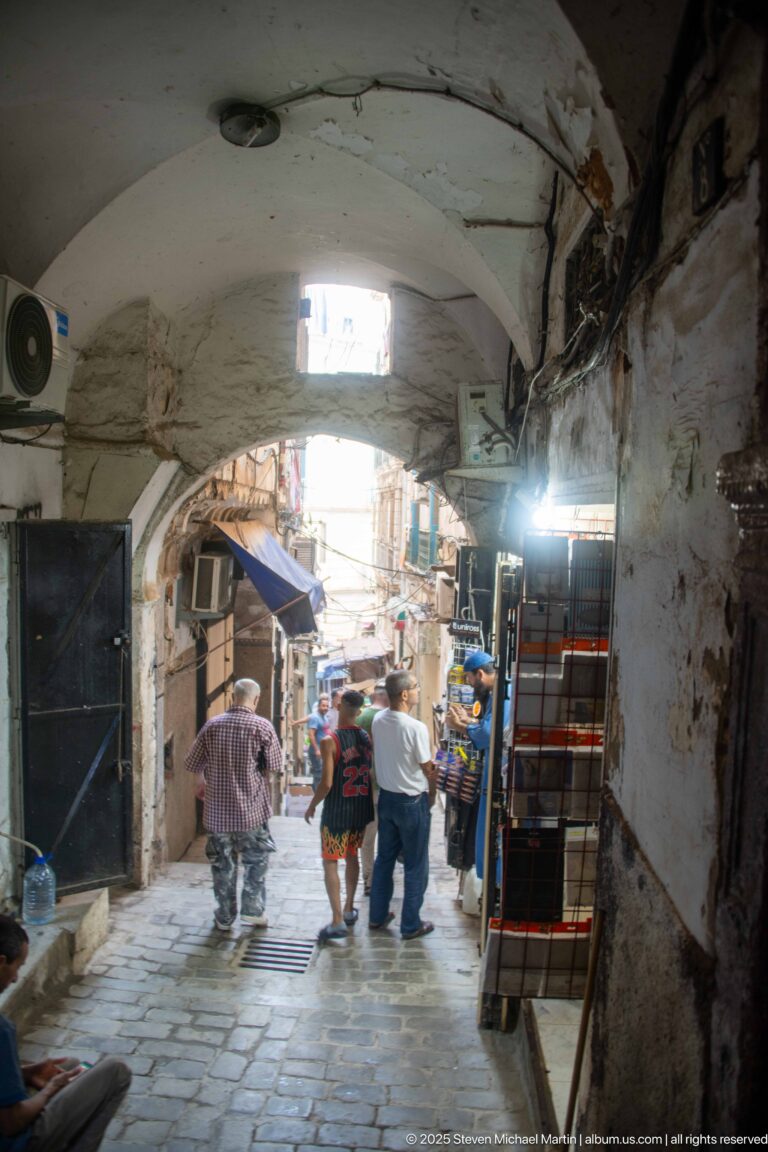 Pedestrians walking through a white arched covered passage toward market stalls and merchandise.