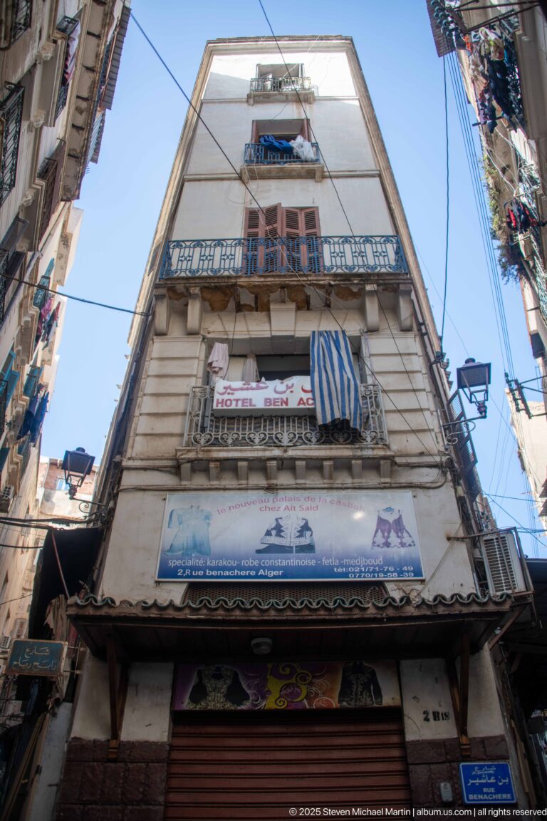 Low-angle view of a tall narrow Casbah building with balconies, shutters, hanging fabric, and signs.