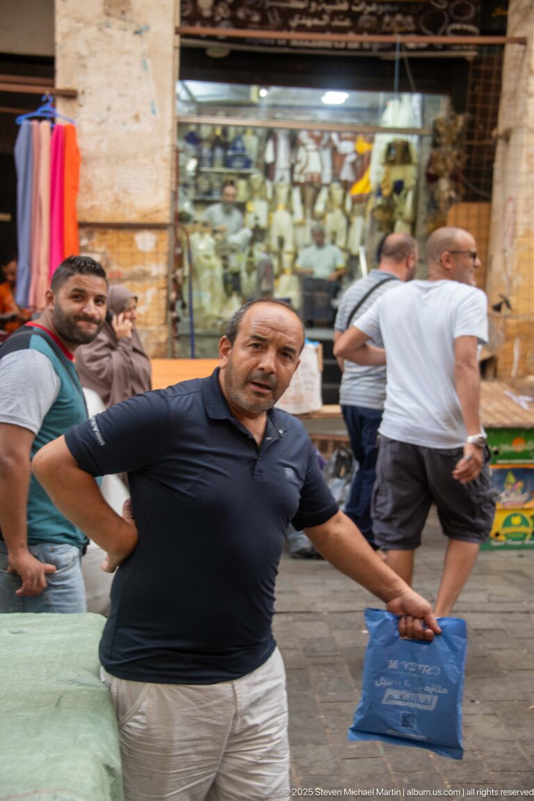 Man in dark polo shirt carrying a blue shopping bag on a crowded Casbah market street.