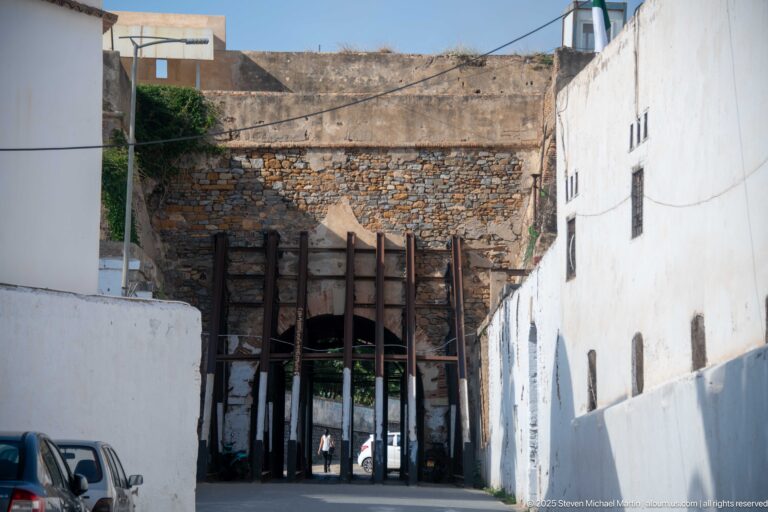 Narrow Casbah street beside a weathered stone wall with a large timber-braced opening, white façades, and overhead wires.