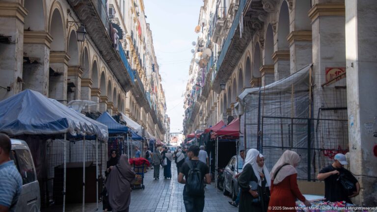 Busy Casbah street lined by repeated stone arches, market canopies, pedestrians, and tall residential façades.