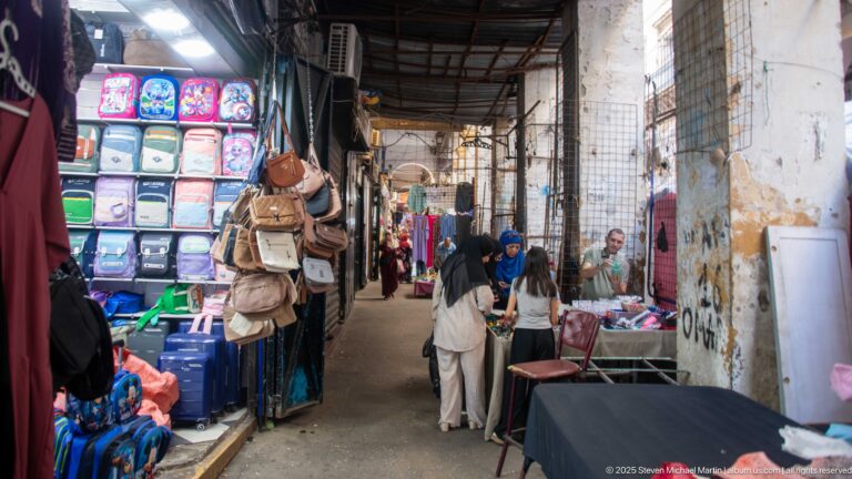 Covered narrow market alley with hanging bags, plastic goods, clothing displays, pedestrians, and weathered walls.