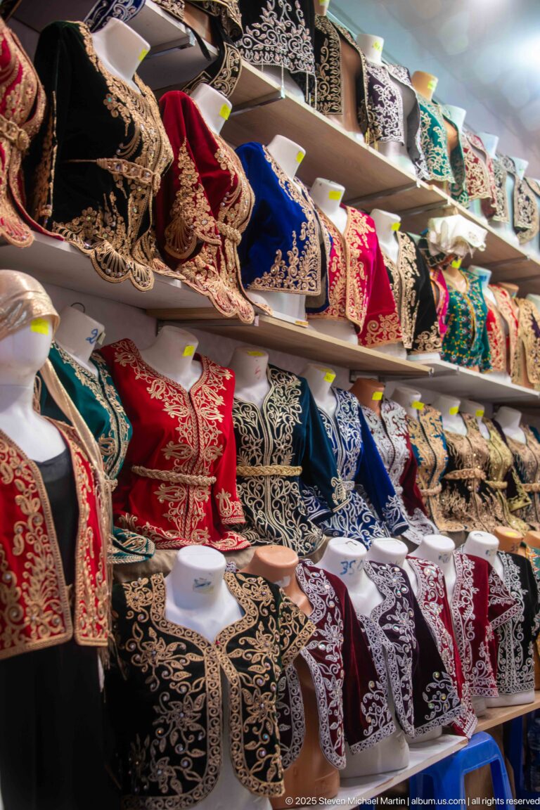 Rows of colorful black, red, blue, and green garments with dense gold and silver embroidery displayed on shop shelves.