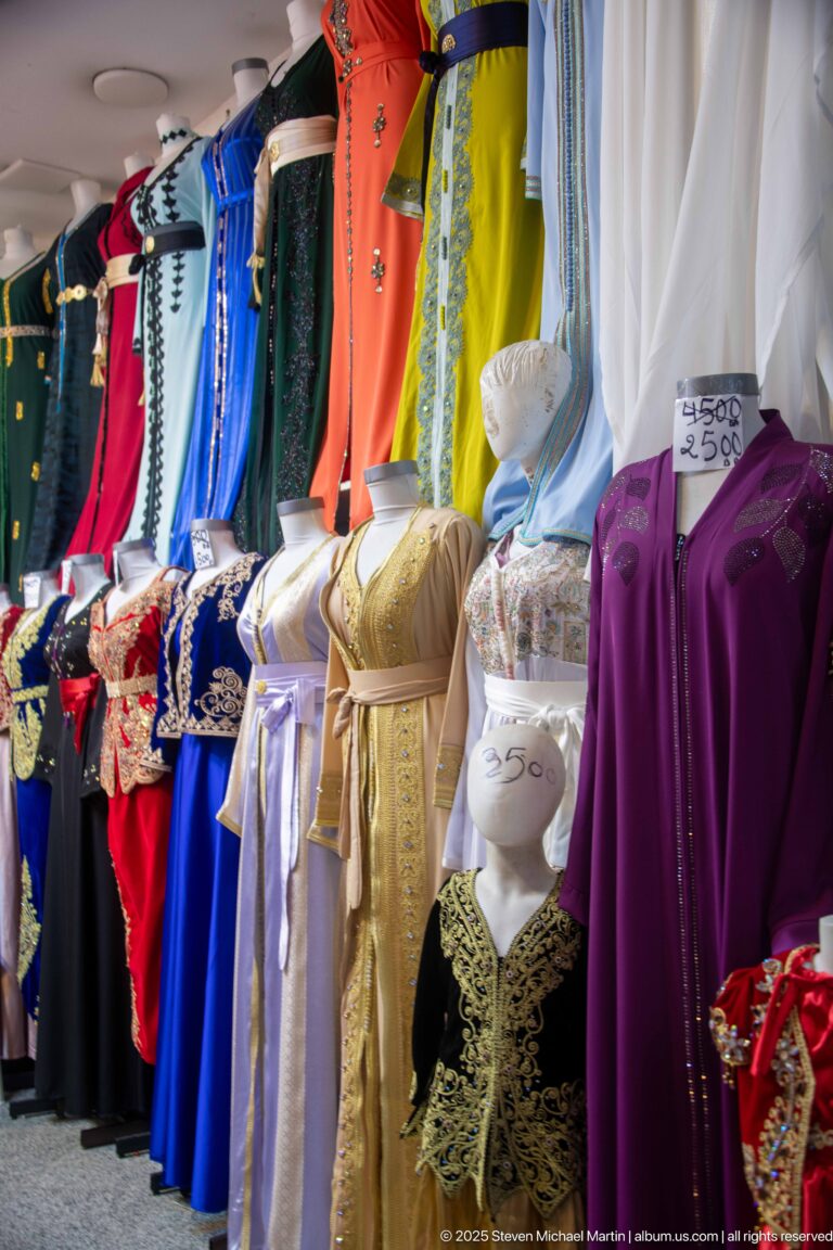 Colorful embroidered women's garments in blue, orange, white, gold, black, and purple displayed with handwritten prices.