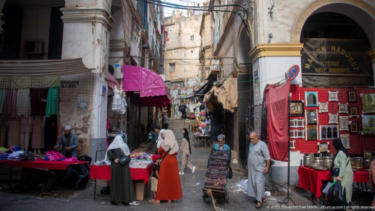 Shoppers and vendors in a narrow Casbah market lane lined with textiles, framed goods, awnings, and weathered buildings.