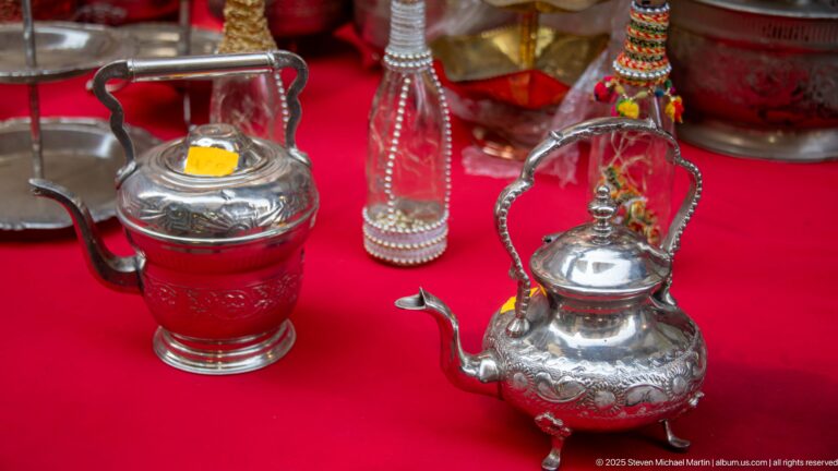 Two ornate silver-colored metal teapots with curved spouts and high handles displayed on bright red cloth.