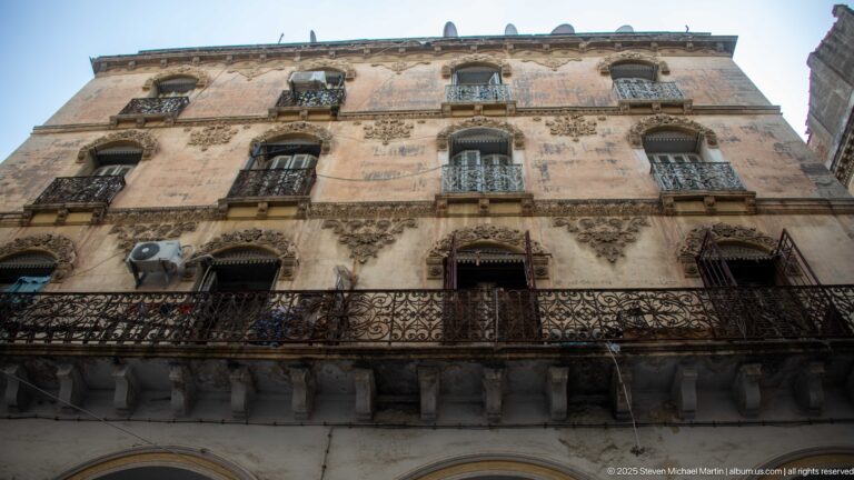 Weathered multi-story Casbah building with ornate window surrounds, wrought-iron balconies, shutters, and air-conditioning units.