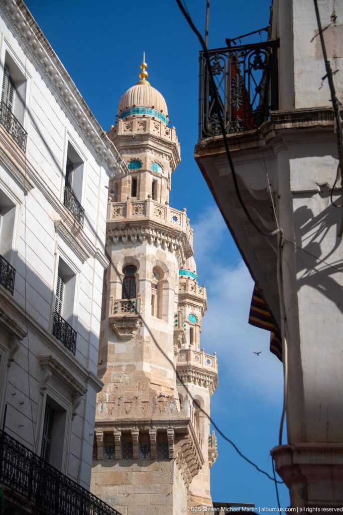 Ornate pale-stone minaret with projecting balconies and turquoise accents rising between white buildings under a blue sky.