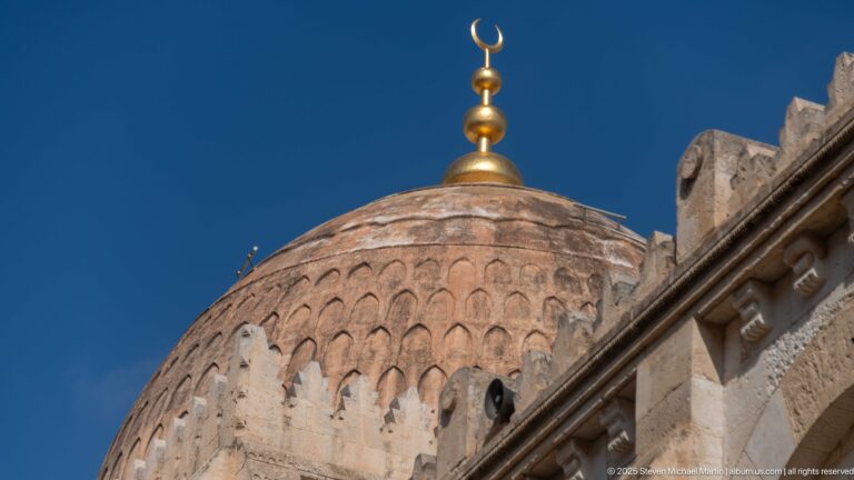 Close view of a patterned masonry mosque dome topped by a gold-colored crescent finial against deep blue sky.