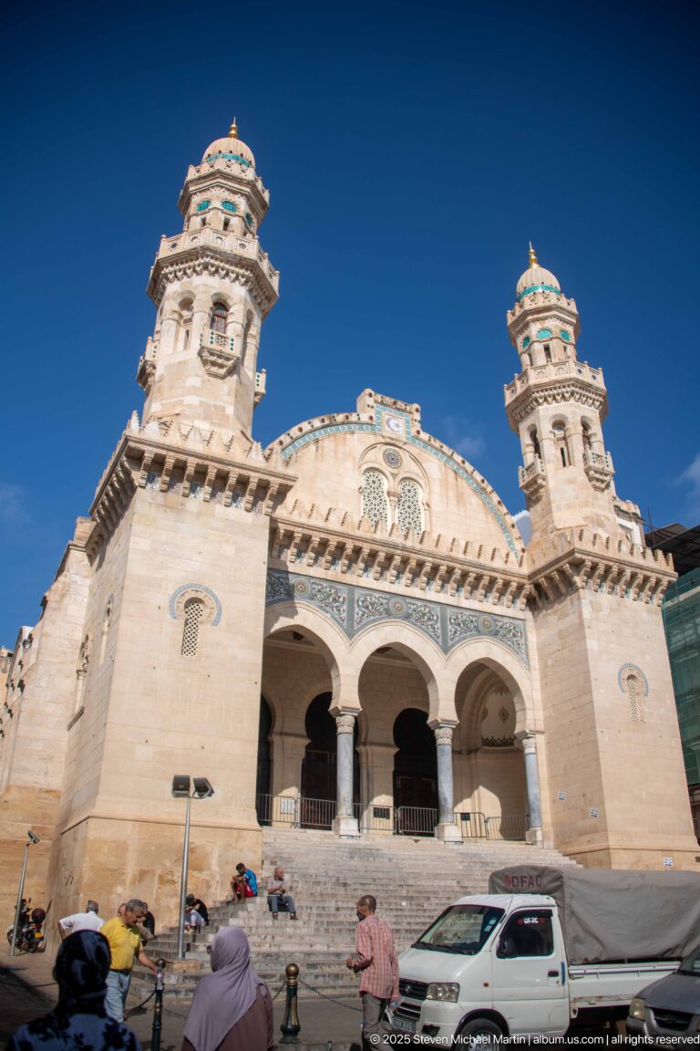 Large pale-stone mosque with twin ornate towers, three entrance arches, broad steps, pedestrians, and vehicles under blue sky.