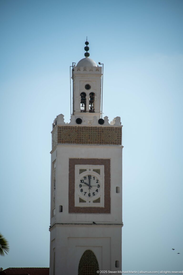 White square clock tower with clock face, red-brown trim, open bell stage, and dark finial against pale blue sky.