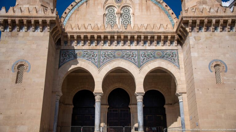 Three tall mosque entrance arches beneath blue-green decorative tile panels, with slender columns and pierced windows above.