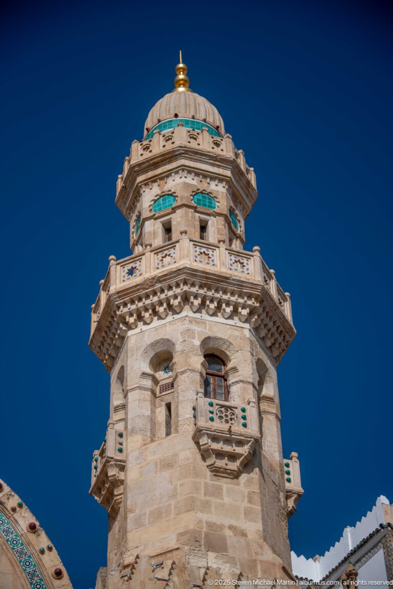 Low-angle close view of an ornate pale-stone mosque tower with carved balconies, turquoise accents, and a ribbed dome.