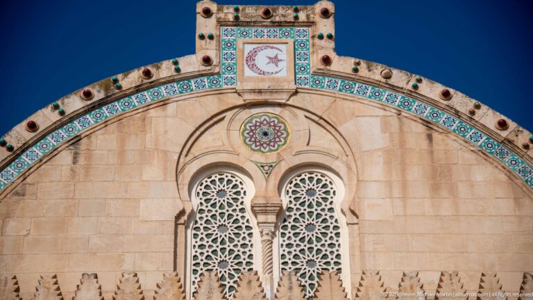 Warm stone mosque gable with paired geometric pierced windows, turquoise tile border, rosettes, and crescent-and-star panel.