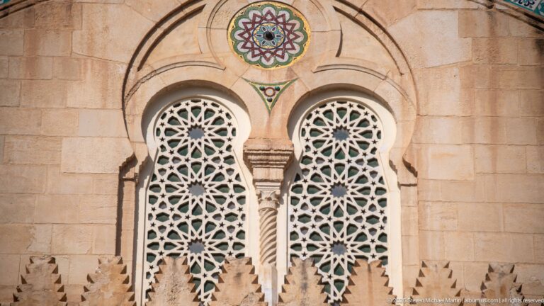 Two tall paired mosque windows filled with white geometric latticework beneath a colorful circular rosette in warm stone.