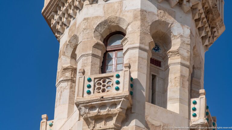 Close view of pale-stone mosque tower with arched openings, projecting carved balcony, and turquoise decorative bosses.