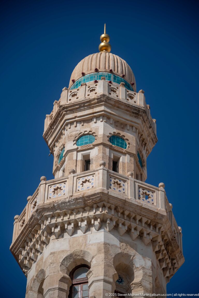 Ornate upper mosque tower with broad carved balcony, turquoise window accents, ribbed dome, and gold-colored finial.
