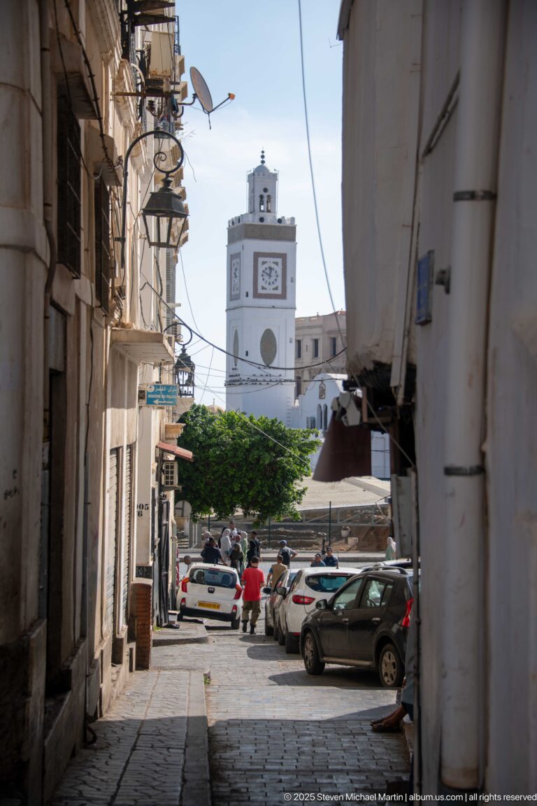 Narrow shaded Casbah street with parked cars leading toward a white clock tower in the sunlit distance.
