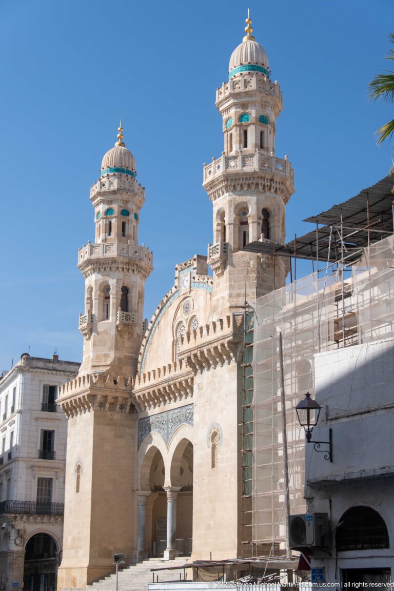 Pale-stone mosque with twin ornate towers and central arches, partly bordered by scaffolding and adjacent city buildings.