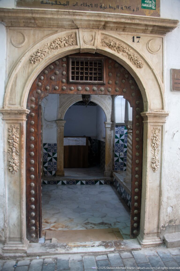 Studded horseshoe-arched doorway with carved stone surround opening into a vestibule with patterned tile walls and columns.