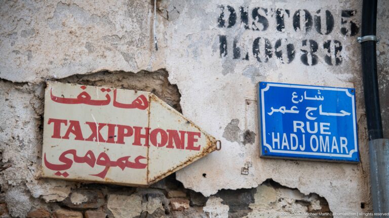 Blue Rue Hadj Omar street plaque beside a worn Taxi-Phone sign with French and Arabic lettering on cracked plaster.