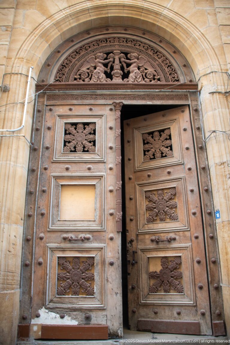 Large carved double wooden doors beneath a stone arch, with floral panels, metal studs, and an ornate semicircular transom.