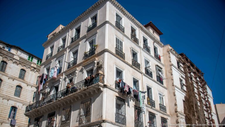 White multi-story corner building with stacked windows, wrought-iron balconies, and laundry hanging beneath a deep blue sky.