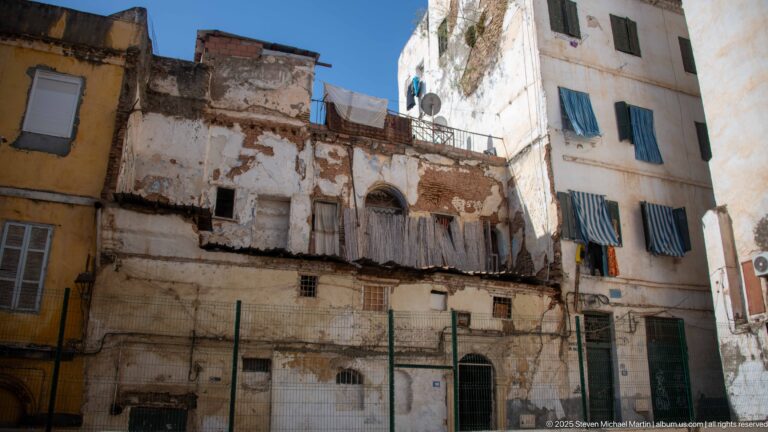 Aging Casbah buildings with peeling plaster, exposed masonry, shuttered windows, laundry, and satellite dishes under blue sky.