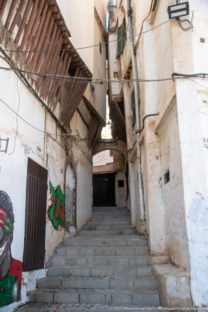 Stone steps climbing a narrow whitewashed Casbah alley beneath projecting wooden structures, wires, shutters, and graffiti.
