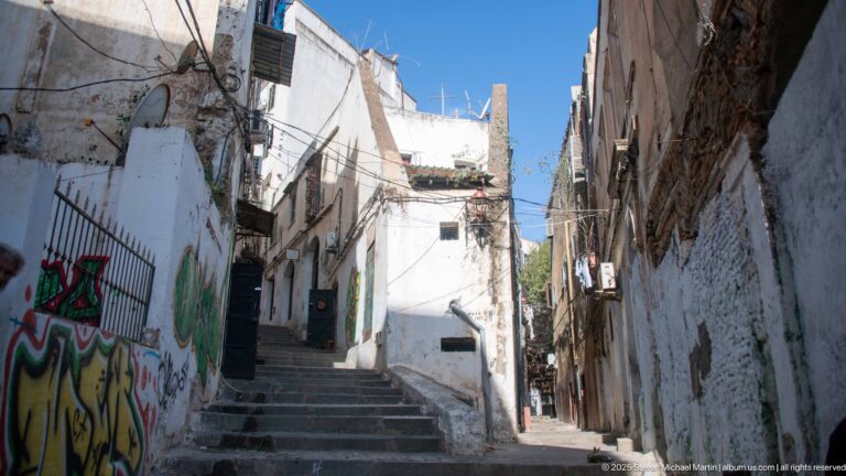 Sunlit stone stairway rising between whitewashed Casbah buildings with graffiti, doorways, utility lines, and blue sky above.