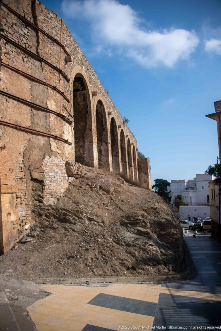 Long weathered masonry wall with a row of tall arched recesses rising beside a street under blue sky.