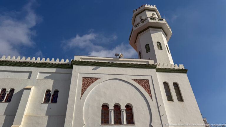White mosque façade with large blind arch, patterned panels, crenellated roofline, and tall angular minaret against blue sky.
