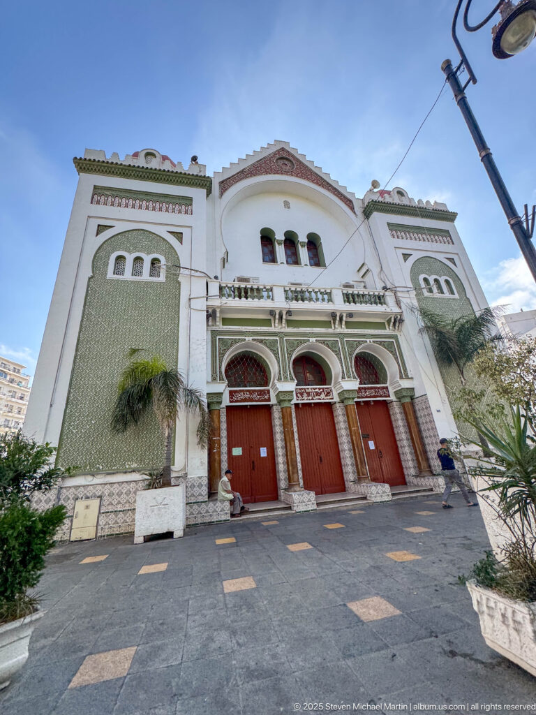 White mosque façade with green geometric decoration, three red doors, arched entrance bays, and paved forecourt.
