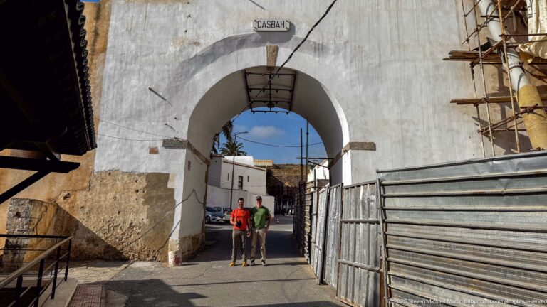 White arched gateway marked CASBAH with two visitors beneath it, construction barriers at the sides, and buildings beyond.