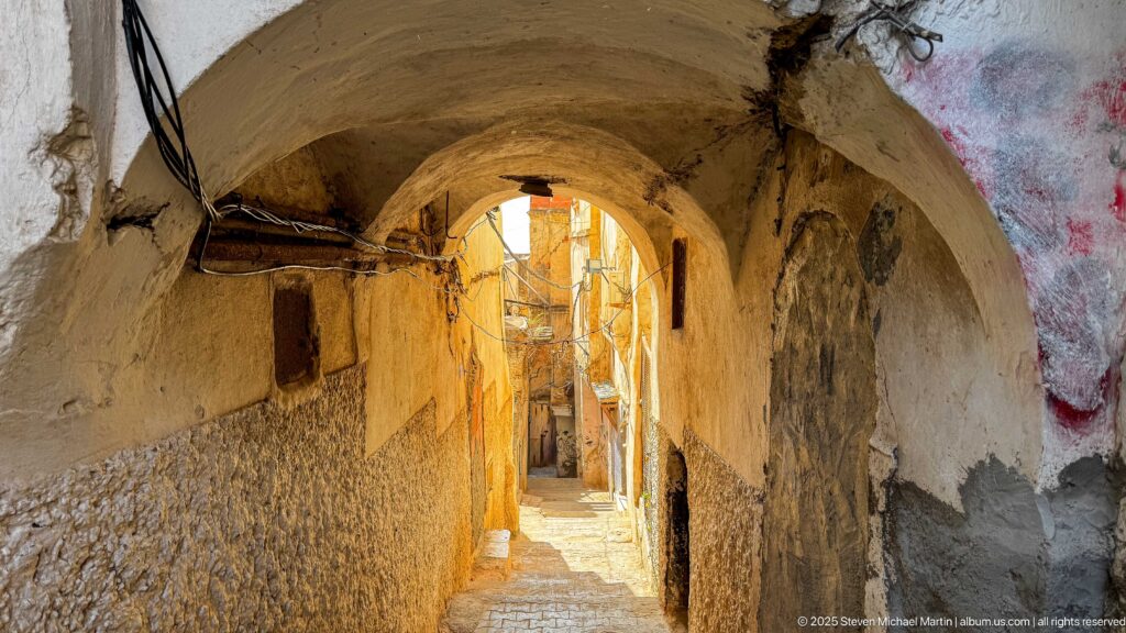 Narrow vaulted Casbah passage with worn plaster walls, stone steps, exposed wiring, and bright sunlight at the far end.