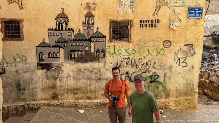 Two visitors standing before a weathered wall with a painted mosque, graffiti, and blue Rue Sidi Ramdane street sign.